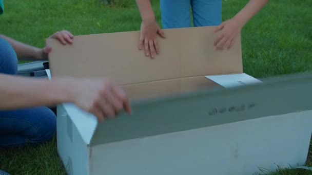 Matka a dvě děti se baví s velkou krabicí na dvorku za slunečného dne. Šťastná rodina si hraje s kartónovou krabicí na trávě. Zpomalený záběr steadicamu. — Stock video