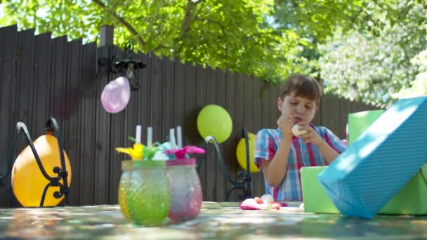 Niño de la escuela tratando de soplar globo para la decoración de la fiesta de cumpleaños. Niño feliz con regalos de color de cumpleaños al aire libre . — Vídeo de stock