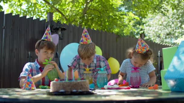 Tres niños felices en sombreros de cumpleaños bebiendo agua de vasos de fiesta de color. Los hermanos celebran la fiesta de cumpleaños al aire libre. Mesa festiva con regalos y tarta . — Vídeos de Stock