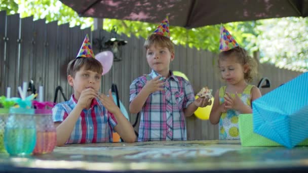 Três crianças felizes em chapéus de festa comendo bolo de aniversário com as mãos ao ar livre. Irmãos e irmã gostando de comer doces no quintal . — Vídeo de Stock