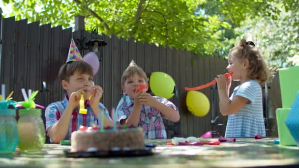 Tre bambini felici in cappelli di compleanno che soffiano tubi di compleanno all'altro all'aperto. Ragazzi e ragazze festeggiano il compleanno con torta e regali. — Video Stock