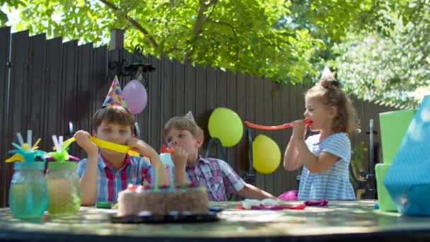 Troje szczęśliwych dzieciaków w urodzinowych kapeluszach dmuchających sobie rury na dworze. Chłopcy i dziewczyna świętują urodziny tortem i prezentami. — Wideo stockowe
