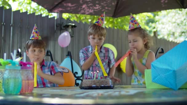 Tres niños felices en sombreros de cumpleaños soplando tubos de cumpleaños el uno al otro al aire libre. Niños y niñas celebran cumpleaños con pastel y regalos . — Vídeos de Stock