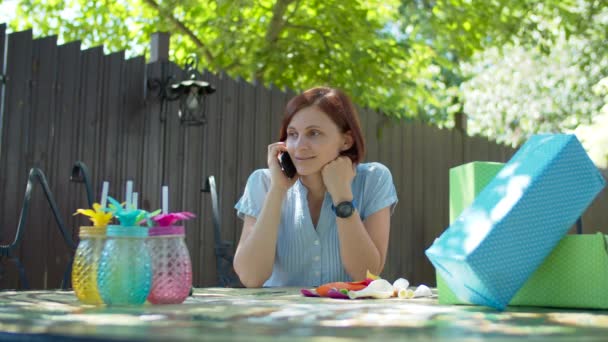 Jovem mulher dos anos 30 conversando por telefone móvel sentado na mesa de aniversário com presentes ao ar livre . — Vídeo de Stock