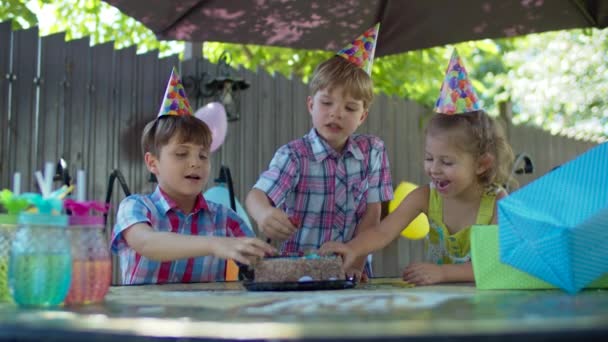Trois enfants heureux dans des chapeaux de fête manger gâteau d'anniversaire avec les mains à l'extérieur. Frères et sœurs appréciant manger des bonbons dans la cour arrière. — Video