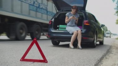 30 'lu yaşlarda bir kadın araba bagajında oturuyor ve otoyoldaki bozuk arabaya yardım etmek için cep telefonu kullanıyor. Kırmızı üçgen, dur işareti. Yolda araba kazası..