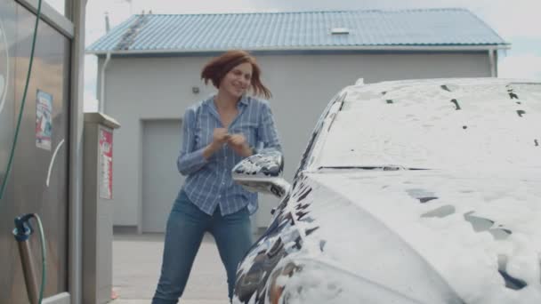 Szczęśliwa kobieta po trzydziestce tańcząca na myjni. Młoda dorosła kobieta chętnie myje samochód pianką i wodą na zewnątrz. — Wideo stockowe