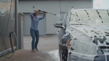 30 'lu yaşlardaki genç bir kadın aile arabasını oto yıkamada yıkıyor. Kadın, dışarıda köpük ve suyla araba yıkar..