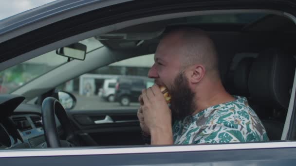 Młody mężczyzna po trzydziestce jedzący hamburgera w samochodzie. Głodny mężczyzna kierowca jedząc lunch fast food na parkingu. — Wideo stockowe