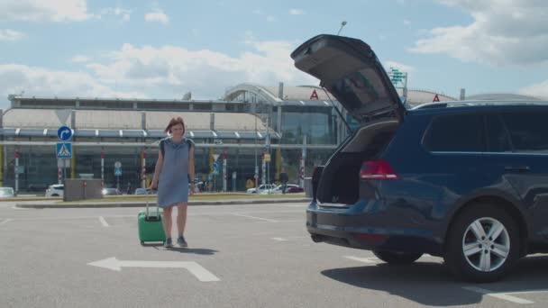 Young 30s woman in dress coming from airport to opened car trunk, putting suitcase and backpack into the car. — Stock Video