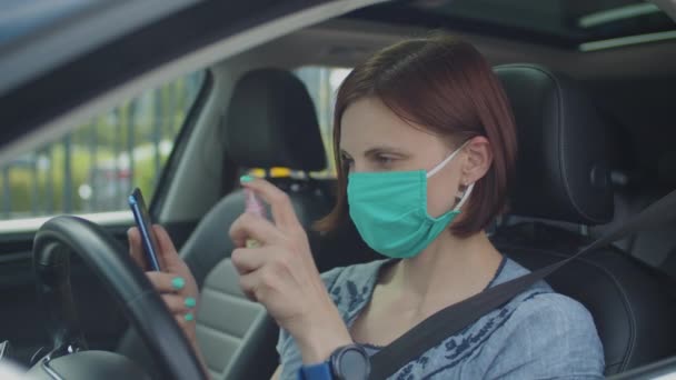 Jovem mulher no rosto máscara protetora usando desinfetante para telefone celular sentado no carro. Motorista usa higienizador para o gadget para prevenir a infecção . — Vídeo de Stock