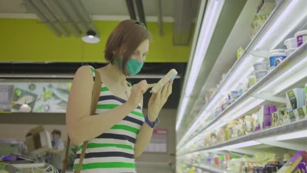 30-jährige Frau mit Gesichtsschutzmaske auf der Suche nach Lebensmitteln im Supermarkt. Frau mit Maske hält ein Stück Käse in den Händen und liest die Zutatenliste. Quarantäne-Einkäufe. — Stockvideo