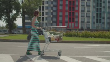 Alışveriş merkezinin yakınındaki otoparkta alışveriş arabasıyla yürüyen genç bir kadın. Market arabası yiyecek ve içecek dolu bir kadın. Yavaş çekim, sabit çekim..