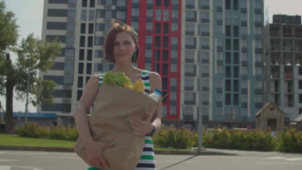 Mujer de 30 años en vestido caminando sosteniendo una gran bolsa de papel en las manos. Mujer con bolsa de comida en el aparcamiento. — Vídeos de Stock
