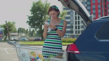 30 'lu yaşlarda bir kadın alışveriş arabasında alışveriş fişi ve masraf takibi ile yemek kontrol ediyor. Kadın sürücü market alışverişini arabanın bagajına koyuyor..
