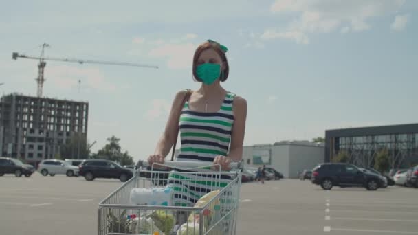 Ung kvinna i ansiktet skyddande mask driver kundvagn på parkering nära köpcentrum. Damen öppnar bagageluckan för dagligvaror. Karantänshopping. Slow motion, steadicam skott. — Stockvideo
