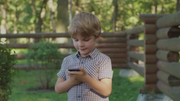 Niño preescolar usando el teléfono móvil sosteniendo la celda en las manos de pie cerca de la valla del parque de madera. Niño con gadget tratando de hacer videollamada fuera. — Vídeo de stock