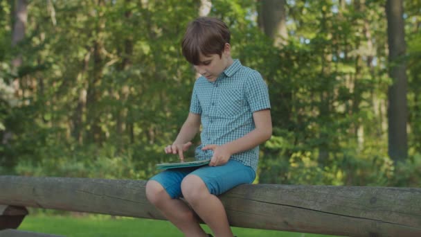 Шкільний хлопчик використовує планшетний комп'ютер, що сидить на паркані. Дитина дивиться гаджет онлайн зовні . — стокове відео
