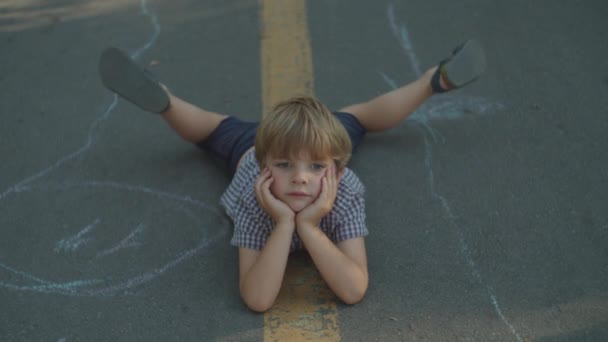 Niño preescolar rubia pensativa que pone en camino en el parque de la ciudad. Niño despreocupado en el suelo afuera. — Vídeo de stock