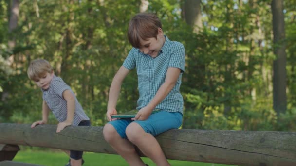 Dua anak dengan satu gadget di luar. Kakak laki-laki menggunakan komputer tablet, adik laki-laki tertarik. Saudara di taman pagar. — Stok Video