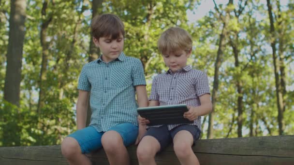Dos niños con un artefacto afuera. El hermano menor que usa la computadora de la tableta, el niño mayor está mirando con interés. Niños en el parque. — Vídeos de Stock