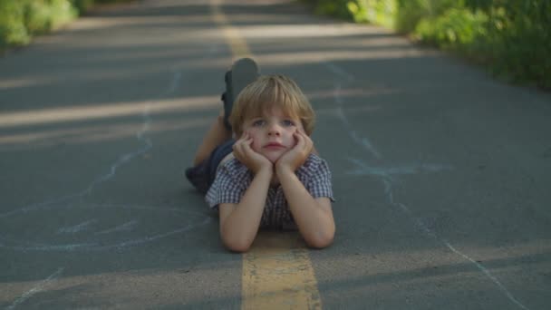 Menino pré-escolar loiro pensativo deitado no caminho no parque da cidade. Miúdo despreocupado no chão lá fora . — Vídeo de Stock