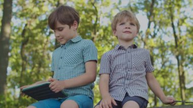 Dışarıda bir aleti olan iki çocuk. Büyük kardeş tablet bilgisayar kullanıyor, küçük kardeş ilgilenmiyor. Çocuk tahta sopayla oynuyor..