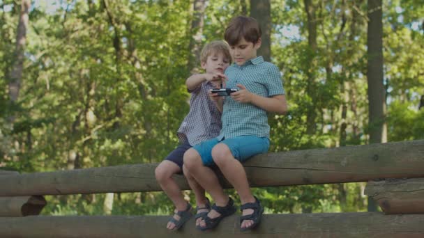 Два мальчика используют мобильный телефон, чтобы смотреть онлайн-контент на сотовом в парке. Дети с гаджетом на улице. — стоковое видео