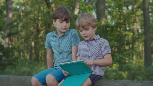Dvě děti s jednou hračkou venku. Mladší bratr používá tablet, starší chlapec se zájmem přihlíží. Děti v parku. — Stock video