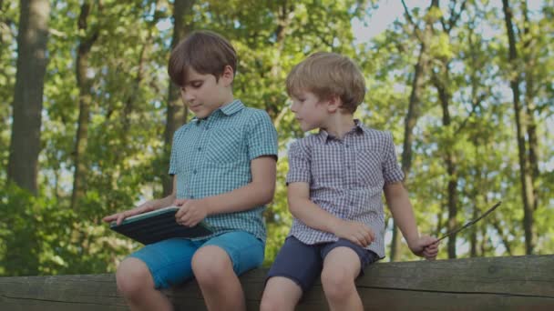 Dvě děti s jednou hračkou venku. Starší bratr pomocí tabletu počítače, mladší bratr se zajímá o bratry online obsahu. — Stock video