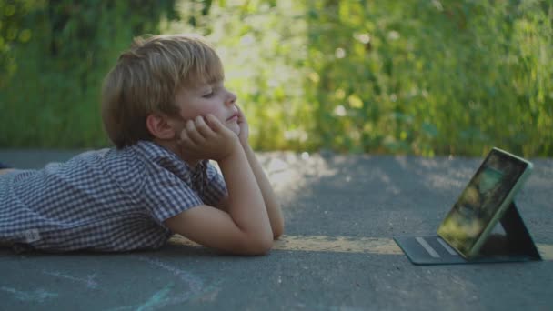 Дошкільний хлопчик використовує планшетний комп'ютер, що лежить на землі в парку. Дитина дивиться гаджет онлайн зовні . — стокове відео