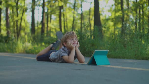 Дошкольник с помощью планшетного компьютера лежит на земле в парке. Ребенок смотрит гаджет онлайн снаружи. — стоковое видео