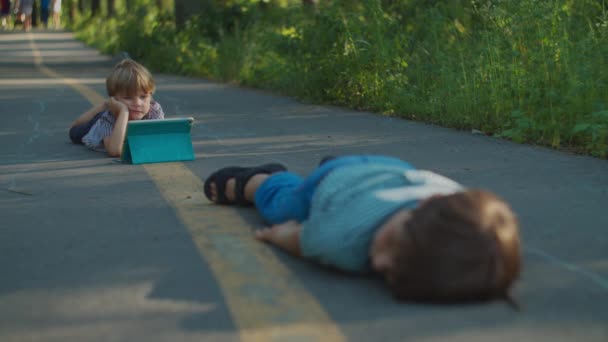 Дошкільний хлопчик використовує планшетний комп'ютер, що лежить на землі в парку. Одна дитина дивиться гаджет онлайн зовні, інша дитина відпочиває на парковій доріжці . — стокове відео