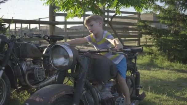 未就学児の少年は外で古いバイクで遊ぶ。子供はオートバイに乗るふりをする。公園内の古代車両. — ストック動画