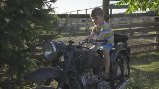 Chłopiec z przedszkola bawiący się starym motocyklem na zewnątrz. Dziecko udaje, że jeździ na motocyklu. Zabytkowy pojazd w parku. — Wideo stockowe