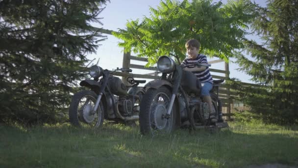 Шкільний хлопчик грає зі старим мотоциклом на вулиці. Дитина робить вигляд, що їде на мотоциклі. Старовинний автомобіль у парку . — стокове відео