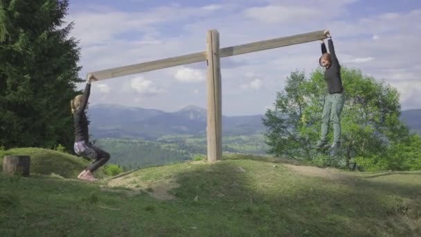 Dwie kobiety jeżdżące na drewnianej huśtawce na szczycie wzgórza z pięknym widokiem na góry. Szczęśliwi przyjaciele cieszą się czasem podczas kempingu. — Wideo stockowe