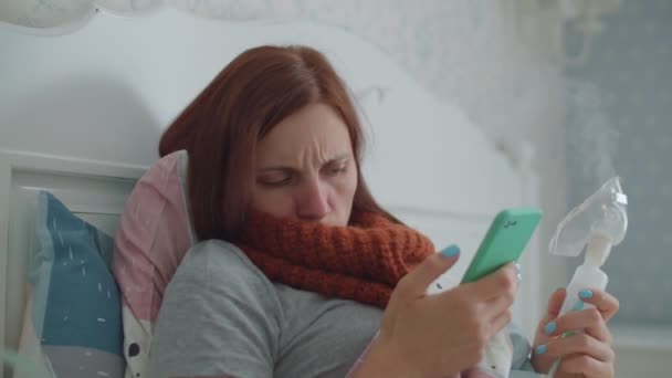 Young 30s woman using inhaler for therapy of disease like flu or respiratory viruses and watching mobile phone at home. Female treating cough with medicine inhaler machine laying in bed. — Stock Video