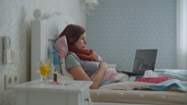 Yatakta dizüstü bilgisayar kullanan hasta bir genç kadın. Boynunda sıcak eşarp olan ve internette doktorla konuşurken burnunu silen bir kadın. Grip ve diğer solunum hastalıkları. Çevrimiçi doktor danışmanlığı.