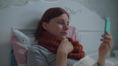 Yatakta cep telefonu kullanan hasta bir genç kadın. Boynunda sıcak eşarp olan ve internette doktorla konuşurken burnunu silen bir kadın. Grip ve diğer solunum hastalıkları. Çevrimiçi doktor danışmanlığı.