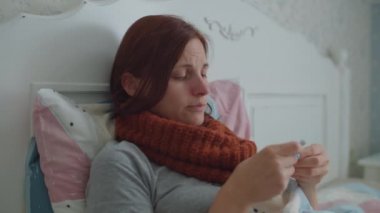 Yatakta uzanmış, öksüren ve ilaçlarını suyla alan, boynunda sıcak atkısı olan hasta genç bir kadın. Grip ve diğer solunum hastalıkları.