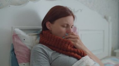 Boynunda sıcak atkısı olan, öksüren ve sıcaklığını elektronik kızılötesi termometreyle ölçen hasta bir genç kadın. Grip ve diğer solunum hastalıkları.