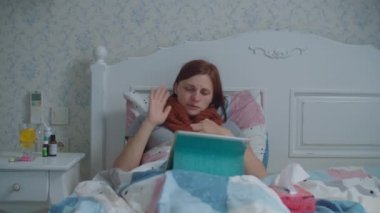 Yatakta tablet bilgisayar kullanan hasta genç bir kadın. Boynunda sıcak eşarp olan ve internette doktorla konuşurken burnunu silen bir kadın. Grip ve diğer virüs hastalıkları. Çevrimiçi doktor danışmanlığı.