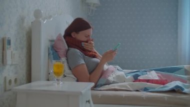 Hasta genç bir kadın elinde cep telefonu ve elinde limonlu bir fincan sıcak çayla yatakta yatıyor. Boyun öksürüğünde sıcak atkısı olan kadın. Grip ve diğer solunum hastalıkları.