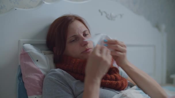 Sick young woman laying in bed. Woman with warm scarf on neck coughs and blows her nose. Hand with cup of hot tea with lemon appears. Virus disease. — Stock Video
