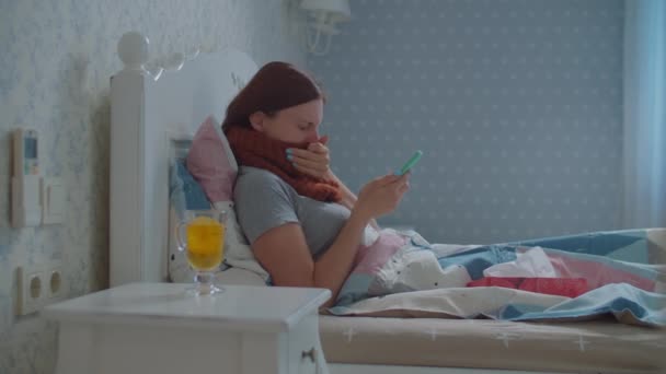 Giovane donna malata sdraiata a letto con telefono cellulare e tazza di tè caldo con limone in mano. Donna con sciarpa calda sul collo tossisce. Influenza e altre malattie respiratorie. — Video Stock