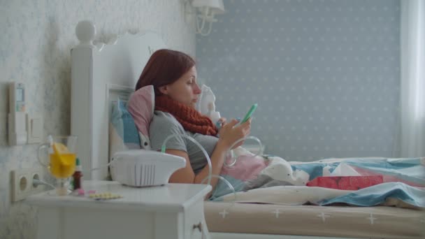 30代の若い女性は、インフルエンザや呼吸器ウイルスなどの病気の治療や自宅で携帯電話を見て吸入器を使用しています。女性の治療咳で薬吸入器マシンはベッドに横たわっています. — ストック動画