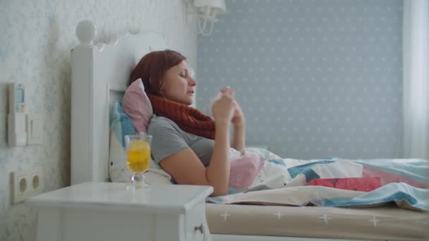 Chorą kobietę z ciepłym szalikiem na szyi leżącą w łóżku i mierzącą temperaturę za pomocą elektronicznego termometru na podczerwień. Grypa i inne choroby układu oddechowego. Choroba z wysoką temperaturą w domu. — Wideo stockowe