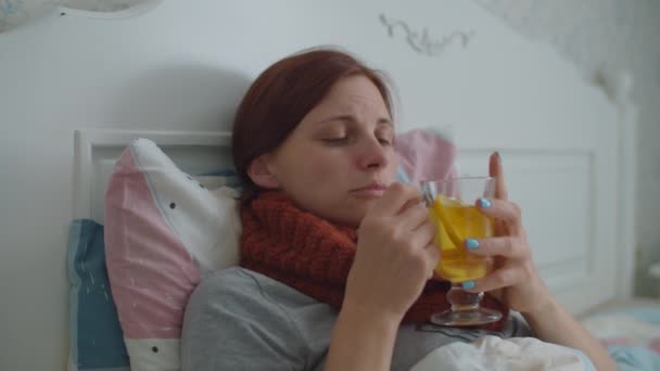 Mujer joven enferma acostada en la cama sosteniendo una taza de té caliente con limón en las manos. Mujer con bufanda caliente en la tos del cuello. Gripe y otras enfermedades respiratorias. — Vídeo de stock