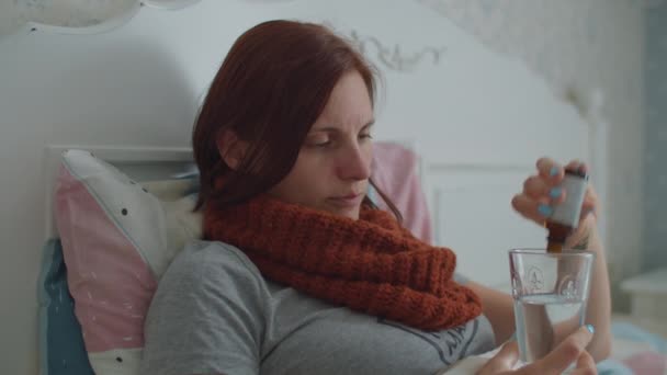 Chora młoda kobieta z ciepłym szalikiem na szyi leżąca w łóżku kaszle i kaszle lekami do wody. Grypa i inne choroby układu oddechowego. — Wideo stockowe
